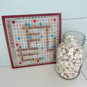 May include: A close-up of a Scrabble game board with tiles spelling out words like "lighthouse", "Egypt", "rock", and "hill". A glass jar filled with white seashells sits next to the board.