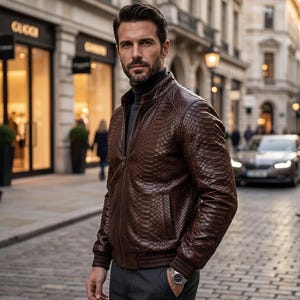 Puede incluir: Una chaqueta bomber de cuero marrón oscuro con un patrón texturizado similar a la piel de serpiente, con cuello alzado, cierre de cremallera frontal y puños acanalados. La chaqueta se lleva sobre un jersey de cuello alto gris oscuro y pantalón gris.