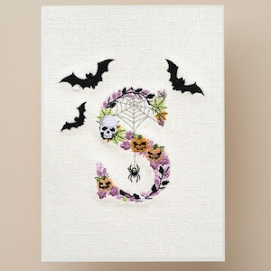 May include: A Halloween-themed embroidered artwork featuring the letter "S". The letter is decorated with a skull, spiderweb, pumpkins, and a spider. Black bats are flying around the letter, creating a spooky and festive design.