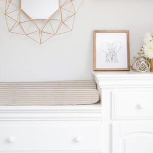 Könnte beinhalten: Weißer Wickeltisch mit einer beige-weißen gestreiften Wickelauflage, einer weißen Schublade und einer weißen Tür. Ein gerahmtes Bild eines Elefanten hängt an der Wand über dem Wickeltisch. Eine Vase mit weißen Blumen steht auf dem Tisch neben dem Bild.