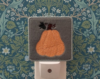 May include: A square night light featuring a gray fabric cover with an embroidered orange pumpkin design. The pumpkin has a dark brown stem and a small black leaf detail. The base of the night light is white plastic with a small circular sensor.