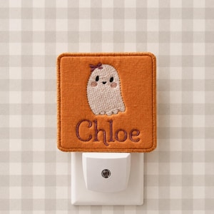 May include: A square orange fabric night light cover featuring an embroidered white ghost with a small purple bow and pink cheeks. The name Chloe is embroidered in purple script below the ghost. The cover is attached to a white plastic plug-in base.