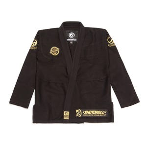 Könnte beinhalten: Eine schwarze Kampfkunst-Gi-Jacke mit goldenen Stickaufnähern und Logos. Auf der Brust befindet sich ein kreisförmiges Shoyoroll-Emblem, während der untere vordere Saum einen rechteckigen Aufnäher mit dem Text Shoyoroll Created for the Superior Grappler aufweist.