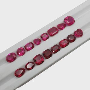 Könnte beinhalten: Eine Sammlung von fünfzehn losen, facettierten Rubinen in verschiedenen Formen wie Oval, Rund, Kissen, Smaragd, Tropfen und Herzschliff. Diese leuchtend tiefroten Edelsteine sind in zwei Reihen auf einem weißen Präsentationstablett angeordnet.