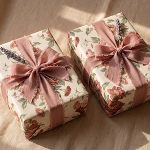 May include: Two rectangular gift boxes wrapped in cream-colored paper featuring a floral pattern of red and pink roses with green leaves. Each box is tied with a wide, frayed-edge dusty rose ribbon and topped with a sprig of dried lavender.