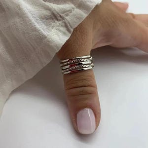 Puede incluir: Un anillo de tono plateado con un diseño de bandas múltiples que alterna texturas lisas y granuladas. El anillo consta de cuatro bandas paralelas distintas que crean una apariencia superpuesta, rodeando el dedo con un acabado metálico pulido.