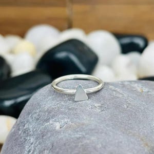 May include: A silver-coloured ring featuring a small, flat, triangular-shaped charm attached to the thin, circular band. The metal surface of the ring has a smooth, polished finish that reflects light, highlighting the geometric shape of the triangle.