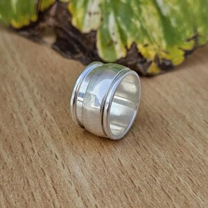 Può includere: Un ampio anello spinner in argento caratterizzato da una fascia centrale liscia e lucida che ruota indipendentemente tra due bordi esterni fissi e più sottili. Il metallo ha una finitura brillante e riflettente con un profilo arrotondato.