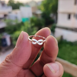 May include: A polished silver ring featuring a prominent infinity symbol design at the center. The band is thin and circular, connecting to the smooth, continuous loops of the infinity emblem. The metal has a bright, reflective finish throughout.