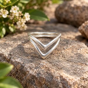 Peut inclure: Une bague en argent poli présentant un design à double bande en forme de V. La bague se compose de deux segments connectés en forme de chevron qui créent une apparence angulaire superposée. La surface métallique est lisse et réfléchissante.