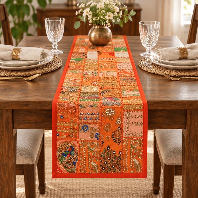 May include: A rectangular table runner featuring a patchwork design of orange fabric squares adorned with intricate embroidery, sequins, and beadwork. The runner displays various patterns including floral motifs and a prominent peacock design.