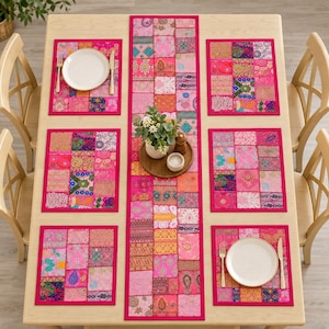Puede incluir: Un juego de mesa de comedor rectangular que incluye un camino de mesa largo de patchwork y cuatro manteles individuales a juego. Cada pieza está hecha de varios cuadrados de tela en tonos rosa con intrincados patrones bordados multicolores y detalles metálicos.