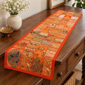 May include: A rectangular orange table runner featuring a patchwork design of embroidered fabric squares. The runner includes intricate beadwork, sequins, and metallic thread detailing, with a prominent peacock motif on one end and a solid orange border.