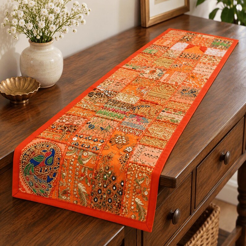 May include: A rectangular orange table runner featuring a patchwork design of embroidered fabric squares. The runner includes intricate beadwork, sequins, and metallic thread detailing, with a prominent peacock motif on one end and a solid orange border.