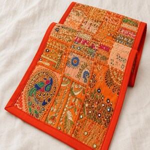 May include: A rectangular table runner featuring a vibrant orange border and a patchwork design of embroidered fabric squares. The panels display intricate gold thread work, colorful floral motifs, and a prominent peacock figure with blue and green accents.