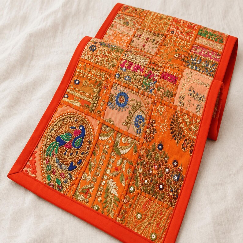 May include: A rectangular table runner featuring a vibrant orange border and a patchwork design of embroidered fabric squares. The panels display intricate gold thread work, colorful floral motifs, and a prominent peacock figure with blue and green accents.