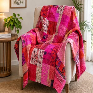 May include: A patchwork quilt featuring a vibrant array of pink, magenta, and red fabric squares with various floral and geometric patterns. The quilt is draped over a light-coloured upholstered armchair, showcasing visible stitching throughout the fabric.