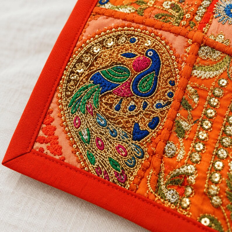 May include: A square textile panel featuring a vibrant orange border and intricate embroidery. The central design showcases a peacock motif in blue, green, and pink threads, accented with gold metallic stitching and small reflective sequins.