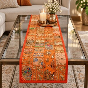 May include: A rectangular table runner featuring a patchwork design of orange fabric squares. Each square is decorated with intricate embroidery, metallic thread, sequins, and small mirrors. The runner has a solid orange border and a peacock motif.