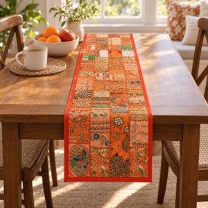 May include: A rectangular table runner featuring a vibrant patchwork design in shades of orange, gold, and red. The runner is decorated with intricate embroidery, small mirrors, and a detailed peacock motif, finished with a solid orange border.