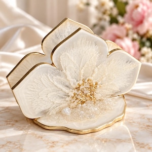 May include: A decorative napkin holder featuring a white resin base with gold-toned edges. The front panel displays a white floral design made of delicate, feathery textures and small, clear, and gold-coloured stone accents embedded in the resin.