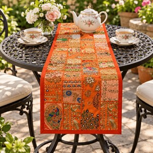 May include: A rectangular orange table runner featuring a patchwork design of various embroidered fabric squares. The runner is decorated with intricate beadwork, sequins, small mirrors, and colorful thread patterns, including a prominent peacock motif.