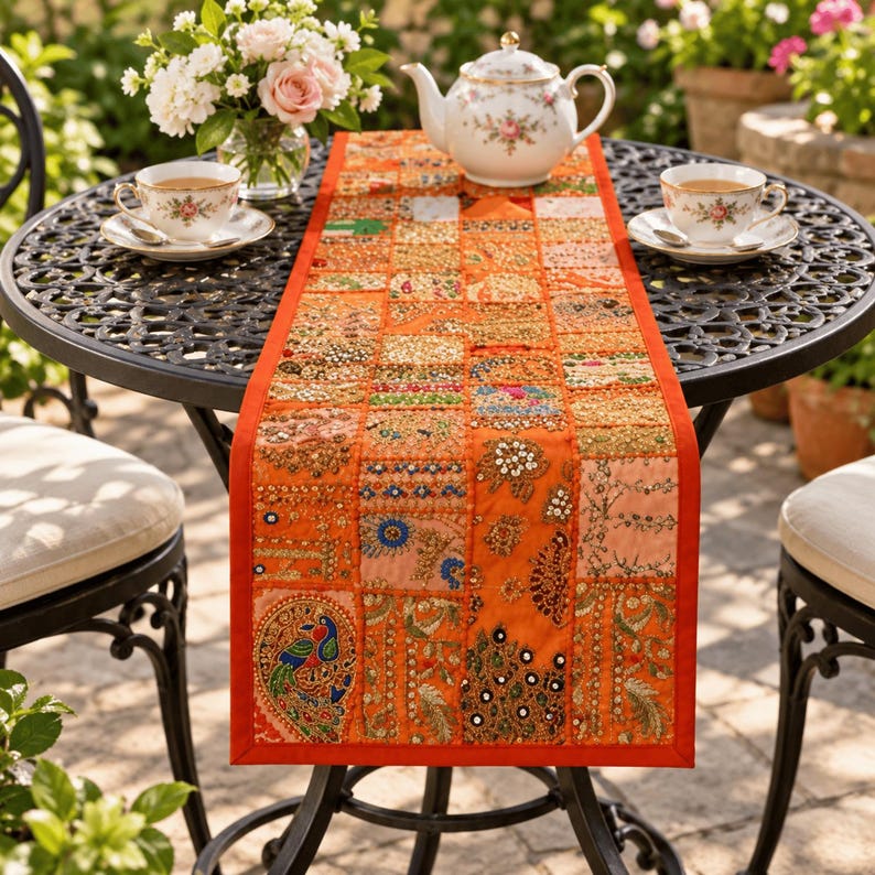 May include: A rectangular orange table runner featuring a patchwork design of various embroidered fabric squares. The runner is decorated with intricate beadwork, sequins, small mirrors, and colorful thread patterns, including a prominent peacock motif.