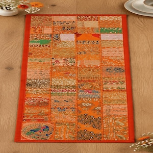 May include: A rectangular table runner featuring a patchwork design of various orange-toned fabric squares. The runner is bordered by a solid orange fabric trim and displays intricate embroidery, sequins, and a prominent peacock motif.