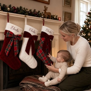 May include: Three Christmas stockings hanging from a mantel, each featuring a white cuff embroidered with the name Elodie. The stockings include a solid red quilted version with a plaid bow, a white quilted version with a red velvet bow, and a red plaid version with a red velvet bow.