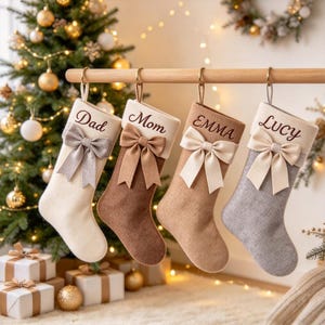 Peut inclure: Quatre chaussettes de Noël personnalisées suspendues à une tringle en bois. Chaque chaussette présente un nœud en tissu et un nom brodé : Dad, Mom, EMMA et Lucy. Elles sont crème, marron, beige et grises, en tissu texturé.