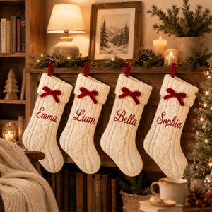 Puede incluir: Cuatro calcetines navideños de punto trenzado en color crema colgados en fila. Cada calcetín tiene un lazo de terciopelo rojo en la parte superior y está bordado con un nombre en letra cursiva roja: Emma, Liam, Bella y Sophia. Hilo grueso y texturizado.