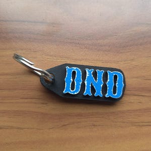 Op de afbeelding: Een zwarte rechthoekige sleutelhanger met afgeronde hoeken voorzien van de verhoogde blauwe tekst DND in een gestileerd lettertype. Een zilveren metalen ring is bevestigd aan de linkerbovenhoek van de sleutelhanger voor gebruik met sleutels.