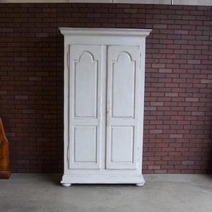 May include: A white, two-door cabinet with a distressed finish. The cabinet has a rectangular shape with a decorative top and base. The doors feature arched panels. The cabinet is set against a brick wall.