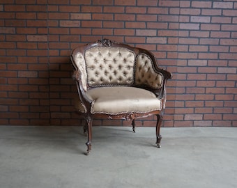 May include: An antique, ornate armchair with a button-tufted back and seat. The chair has a dark wood frame with carved details and a light-coloured fabric. The chair is set against a brick wall.