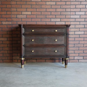 May include: A dark brown wooden dresser with three drawers and gold-colored knobs. The dresser has gold accents on the legs and is set against a brick wall background. The dresser is approximately 36 inches wide.