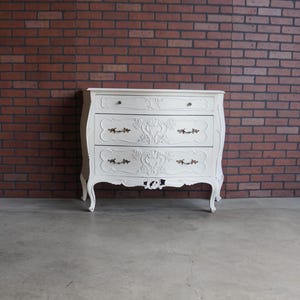 May include: A white, ornate dresser with three drawers and decorative hardware. The dresser has curved legs and detailed carvings on the drawer fronts. The background is a brick wall.