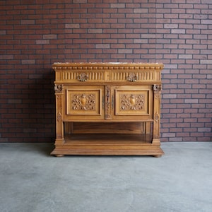 May include: A vintage wooden cabinet with intricate carvings and a marble top. The cabinet has two doors with ornate details and a lower shelf. The cabinet is standing on a concrete floor in front of a brick wall.