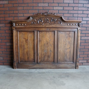 May include: A dark brown, antique wooden headboard with ornate carvings. The headboard features a decorative top with floral and scroll details. The wooden panels are vertically oriented and have a rich, aged appearance. The headboard is against a brick wall.