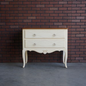 May include: A white two-drawer dresser with ornate legs and decorative hardware. The dresser has a wood top and is in front of a brick wall.