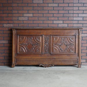 May include: A large, antique wooden headboard with intricate floral carvings. The headboard is a warm brown color and features detailed relief carvings. It stands against a brick wall.