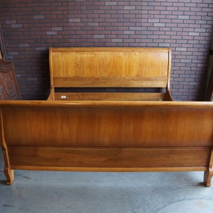 May include: A light brown wooden sleigh bed frame featuring a tall, curved headboard and a matching lower footboard with smooth, polished surfaces and visible wood grain patterns, designed with a classic scroll-top silhouette.