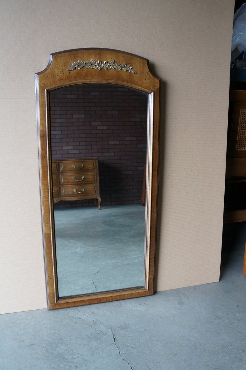 K&ouml;nnte beinhalten: Ein hoher, mit Holz gerahmter Spiegel mit einem Blattgold-Design an der Oberseite. Der Spiegel hat eine abgeschr&auml;gte Kante und spiegelt eine Kommode mit drei Schubladen wider.