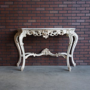May include: White painted console table with ornate carvings and a marble top. The table has a traditional design with curved legs and a detailed base.