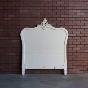 May include: White wooden headboard with ornate carvings and a curved top. The headboard is against a brick wall.