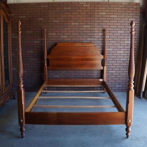 May include: A wooden four-poster bed frame with a rich brown finish. The bed features a tall headboard with a curved top edge and four tall, spiraled posts topped with finials. The frame includes side rails and wooden slats for mattress support.