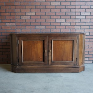 May include: A dark brown wooden cabinet with two doors and brass knobs. The cabinet has a slightly curved front and sits on a base with a decorative trim.
