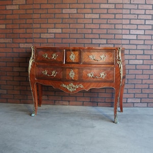 May include: A wooden dresser with four drawers and ornate brass hardware. The dresser has a curved front and legs, with a reddish-brown finish and decorative gold accents. The background is a brick wall.
