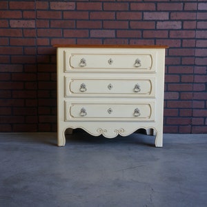 May include: A cream-colored wooden chest of drawers featuring three horizontal drawers with decorative molding, silver-toned ring pulls, and keyhole escutcheons. The piece has a natural wood-toned top surface and a scalloped apron base design.
