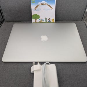Könnte beinhalten: Ein silberner Laptop mit geschlossenem Deckel und Apple-Logo, begleitet von einem weißen Netzteil mit aufgewickeltem Kabel. Hinter dem Laptop befindet sich eine rechteckige Karte mit blauem Himmel, einem grünen Baum, Zeichentrickfiguren und dem Text Chaos Collectables.