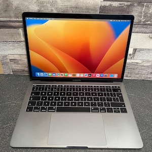 Puede incluir: Un ordenador portátil MacBook Pro de color plateado con teclado negro y un trackpad grande. La pantalla muestra un fondo de pantalla abstracto en tonos naranja y azul vibrantes, con un dock de aplicaciones en la parte inferior y una barra de menú.